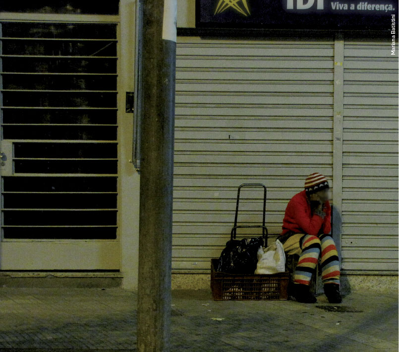 Foto de uma mulher. É noite e ela está sentada na rua, com alguns de seus pertences - sacolas - em uma caixa. No topo da imagem, em um placa de loja está a frase "Viva a diferença"