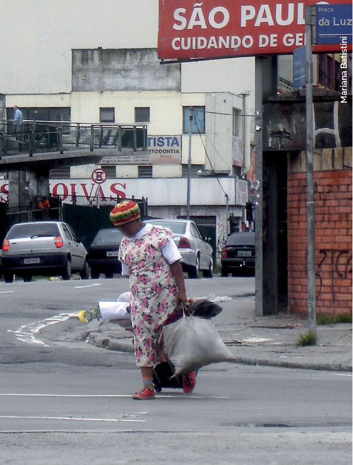 Fotografia de uma mulher no centro de São Paulo. Ela atravessa a rua com algumas sacolas e uma mala. Na imagem também aparece uma placa com os dizeres "São Paulo cuidando da gente"