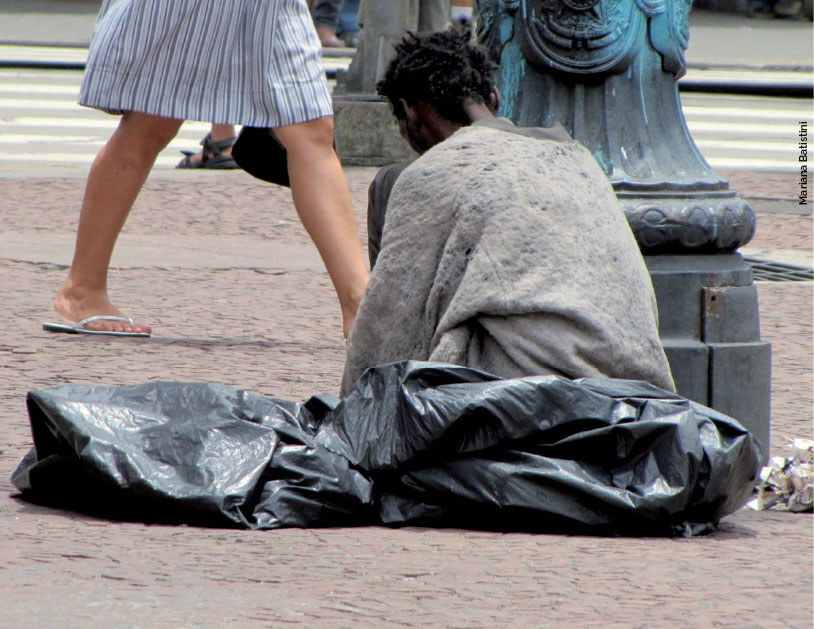 Uma mulher está sentada na rua, entre sacos de lixo, enrolada em um cobertor velho.