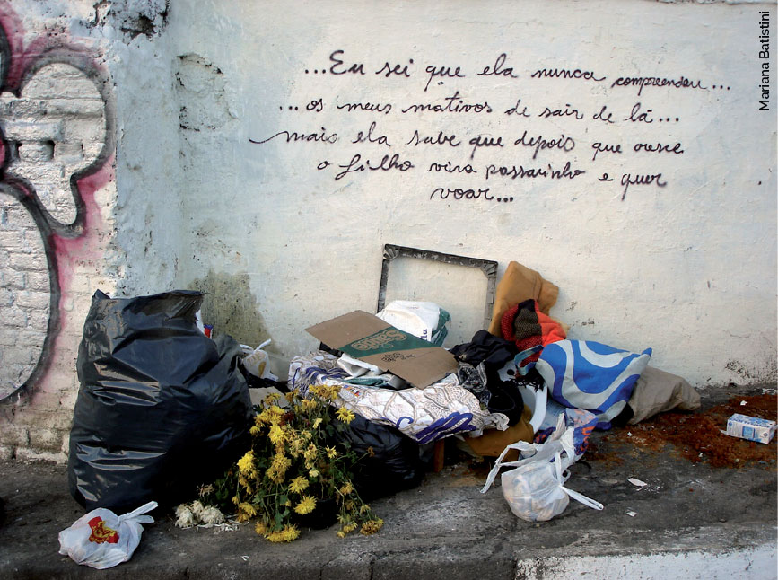 Alguns itens - sacolas, flores, roupas - estão na calçada; atrás deles, em uma parede, está escrito "Eu sei que ela nunca compreendeu... os meus motivos de sair de lá... 'mais' ela sabe que depois que cresce o filho vira passarinho e quer voar...