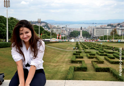 Fotografia de Amanda em Portugal, ela está sentada em um muro com uma paisegem de jardim e cidade ao fundo