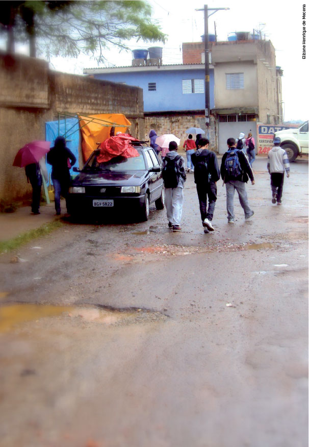 Em uma rua esburacada, crianças voltam da escola - elas tem mochilas nas costas. O dia está cinza e chuvoso.