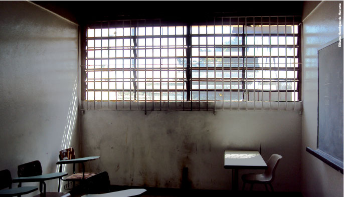 fotografia de uma sala de aula, as paredes tem marcas de fuligem, o sol entre por uma janela, mas ela está cheia de grades