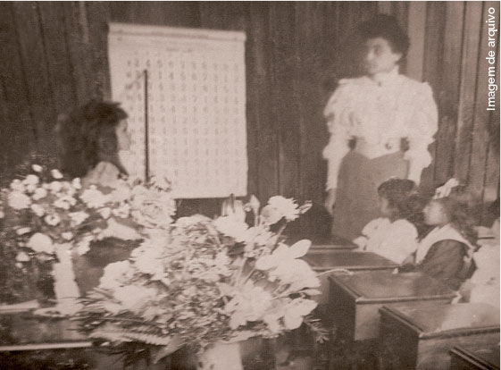 Fotografia antiga, professora e alunas estão com roupas de época em uma sala de aula