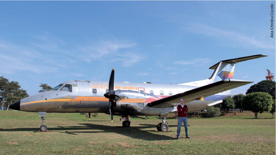 Milioni, em frente a um avião. Ele está em um gramado e o dia está muito ensolarado, com o céu azul