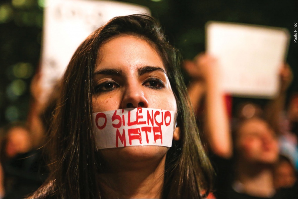 Mulher em um protesto, com uma fita na boca escrito - O silêncio mata