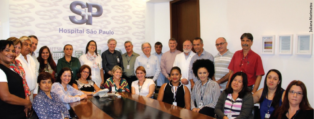 Equipe reunida ao redor de uma mesa, ao fundo o símbolo do Hospital São Paulo