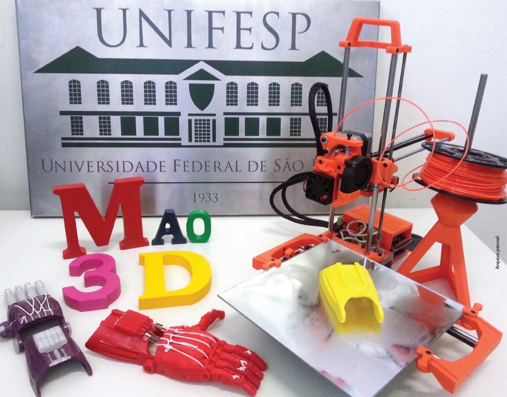 Em uma mesa estão próteses de mão, equipamento de impressão, uma placa com a marca Unifesp e o texto impresso Mão 3D