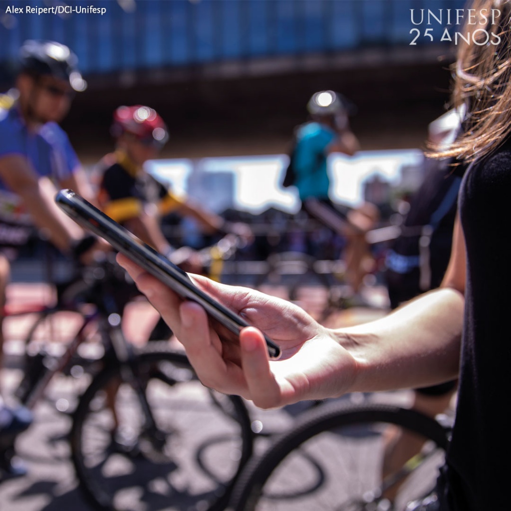 Imagem de uma mulher segurando um celular em primeiro plano, e ao fundo, pessoas andando de bicicleta