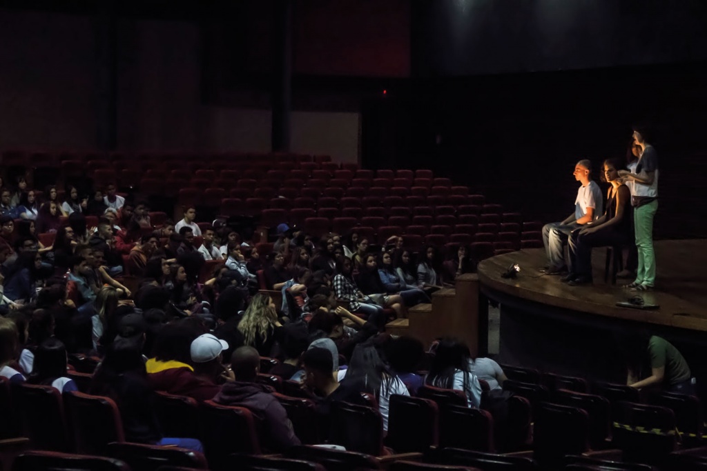 Imagem de um teatro, há atores no palco e pessoas na plateia