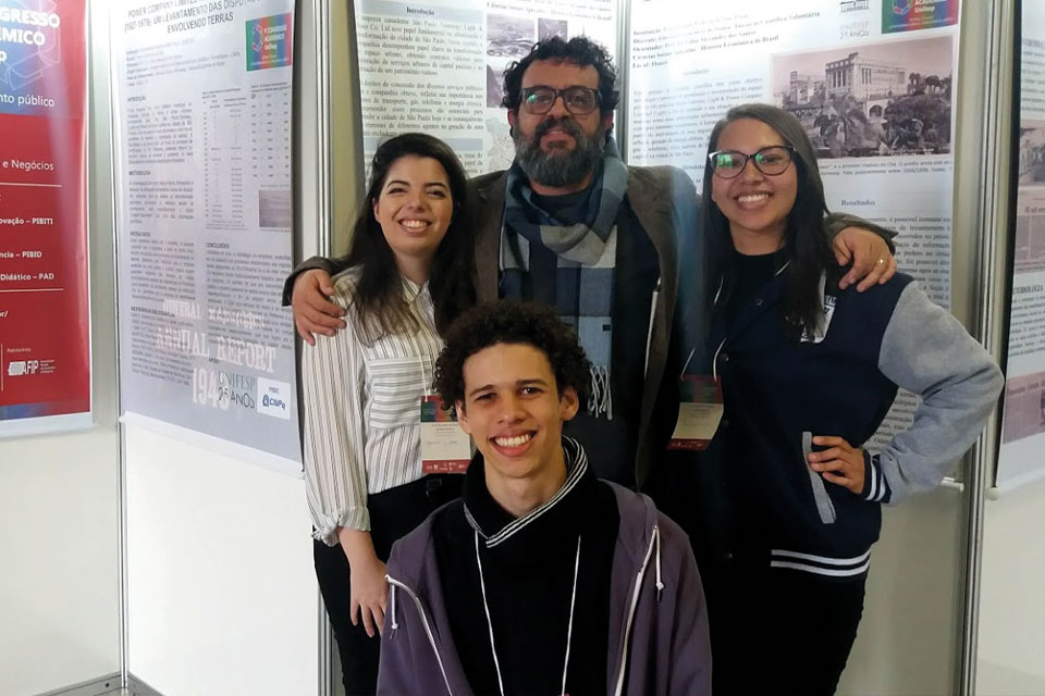 Professor e três jovens em frente a banners acadêmicos