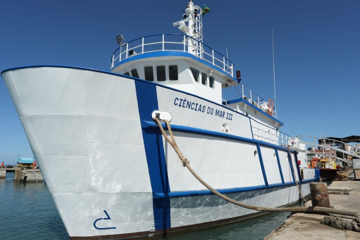 Navio está ancorado. Ele é branco com detalhes em azul. Nele, lê-se o nome do projeto Ciências do Mar III