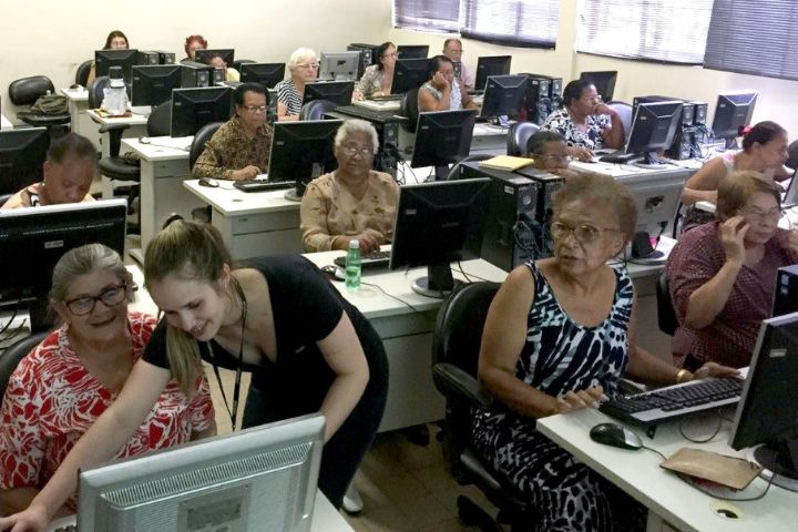 Senhoras em uma sala de aula utilizando computadores; em primeiro plano, mulher auxilia uma aluna