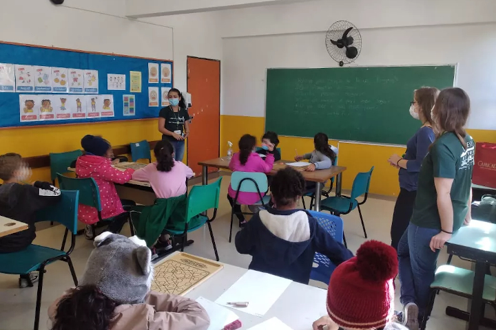 Sala de aula da escola Paulistinha, bem colorida, com as crianças sentadas na carteira prestando atenção na explicação das graduandas do PET Saúde.