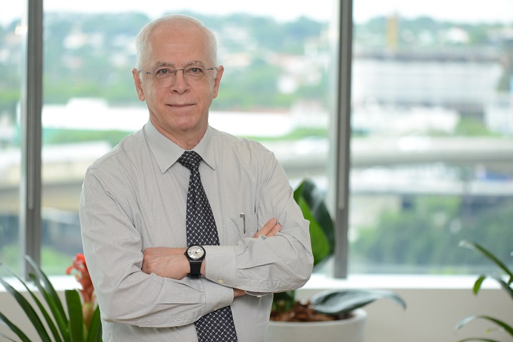 Médico grisalho e de óculos, com camisa social branca e gravata preta, cruzando os braços