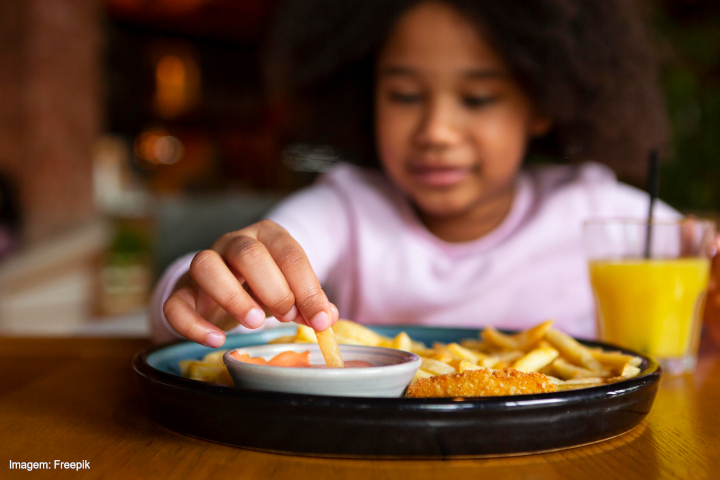 A imagem mostra uma criança sentada à mesa, pegando um pedaço de batata frita de um prato. O prato contém batatas fritas, nuggets e um molho rosado em um recipiente. Ao fundo, há um ambiente desfocado, indicando um possível restaurante ou lanchonete. À direita do prato, há um copo com suco de laranja. A criança está vestida com uma blusa clara e tem cabelo volumoso e encaracolado. A cena sugere um momento de refeição descontraído.   Legenda no canto inferior esquerdo: "Imagem: Freepik".