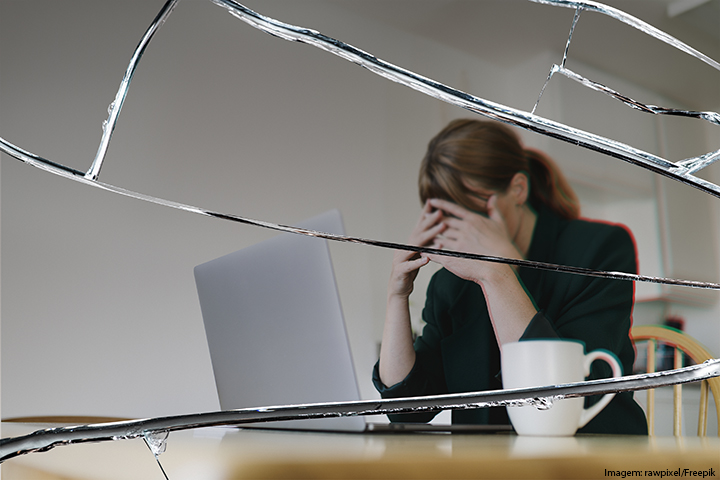 Mulher em uma mesa com computador com as mãos no rosto; em primeiro plano, aparece um vidro quebrado