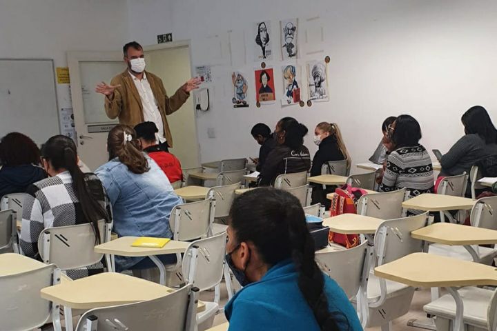 Professor dando aula em pé e com máscara; alunos sentados com máscara 