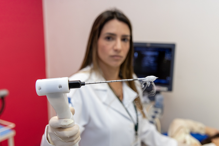 A imagem mostra uma profissional de saúde, usando jaleco branco e luvas, segurando um dispositivo médico avançado, que parece ser uma agulha conectada a um aplicador utilizado para procedimentos minimamente invasivos. A ponta da agulha está envolta por uma névoa ou vapor, possivelmente indicando um processo de resfriamento, como ocorre na crioablação. Ao fundo, desfocado, há um monitor de ultrassom e parte de uma paciente deitada, sugerindo um ambiente clínico ou hospitalar. A expressão séria da profissional e o foco no dispositivo destacam a importância e a precisão do procedimento.