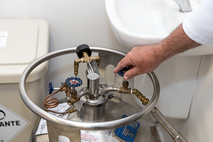 A imagem mostra a mão de um homem, manipulando um cilindro metálico em um ambiente que parece ser um laboratório. O cilindro possui válvulas e mangueiras conectadas a ele, sugerindo que contenha algum tipo de gás ou líquido sob pressão. O homem está segurando uma das válvulas com a mão, indicando que está realizando algum tipo de ajuste ou manobra no equipamento. Ao lado do cilindro, há uma pia com torneira e um recipiente com a inscrição "ANTE", que pode ser um recipiente para descarte de materiais contaminados. A imagem transmite a ideia de um procedimento técnico e cuidadoso, realizado em um ambiente controlado. O jaleco branco do homem reforça a ideia de um profissional da área da saúde ou de um laboratório, enquanto os equipamentos e os símbolos de segurança presentes na imagem sugerem a manipulação de substâncias potencialmente perigosas.