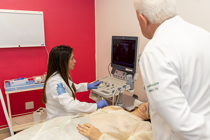 A imagem mostra uma cena médica em que dois profissionais de saúde, um homem de cabelos brancos e uma mulher de cabelos castanhos, estão realizando um exame ou procedimento em uma paciente. A mulher opera um aparelho de ultrassom, com a tela mostrando uma imagem médica, enquanto o homem observa. A paciente está deitada em uma maca, parcialmente coberta por um lençol, com parte do tórax exposto para o exame. A sala tem paredes com cores claras, incluindo um painel vermelho ao fundo, e equipamentos médicos estão dispostos no ambiente.